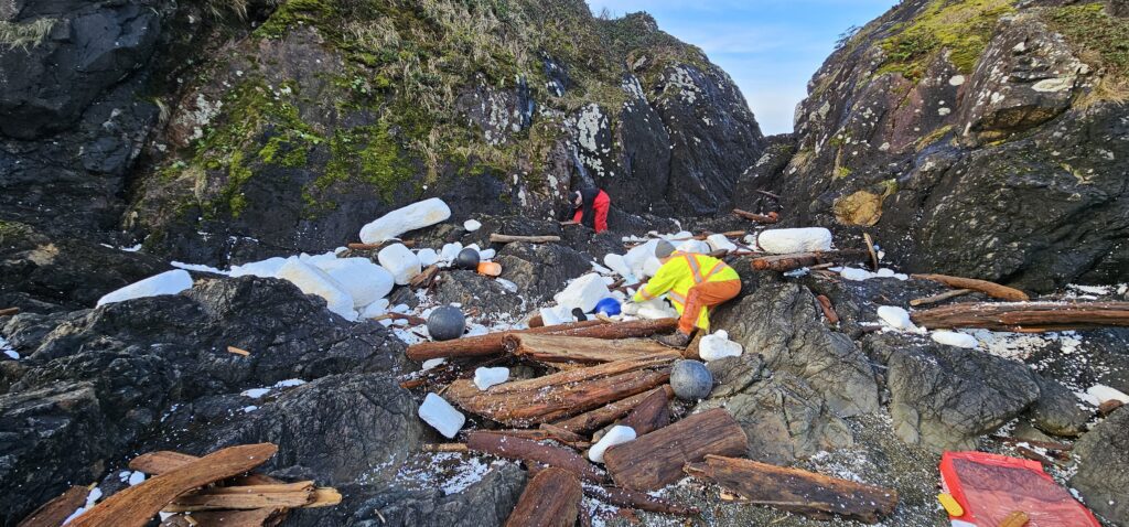 Ocean Legacy team cleaning a remote beach with plastics and large styrofoam pieces, driftwood, rocks, and people wearing high vis clothing bending over collecting plastic