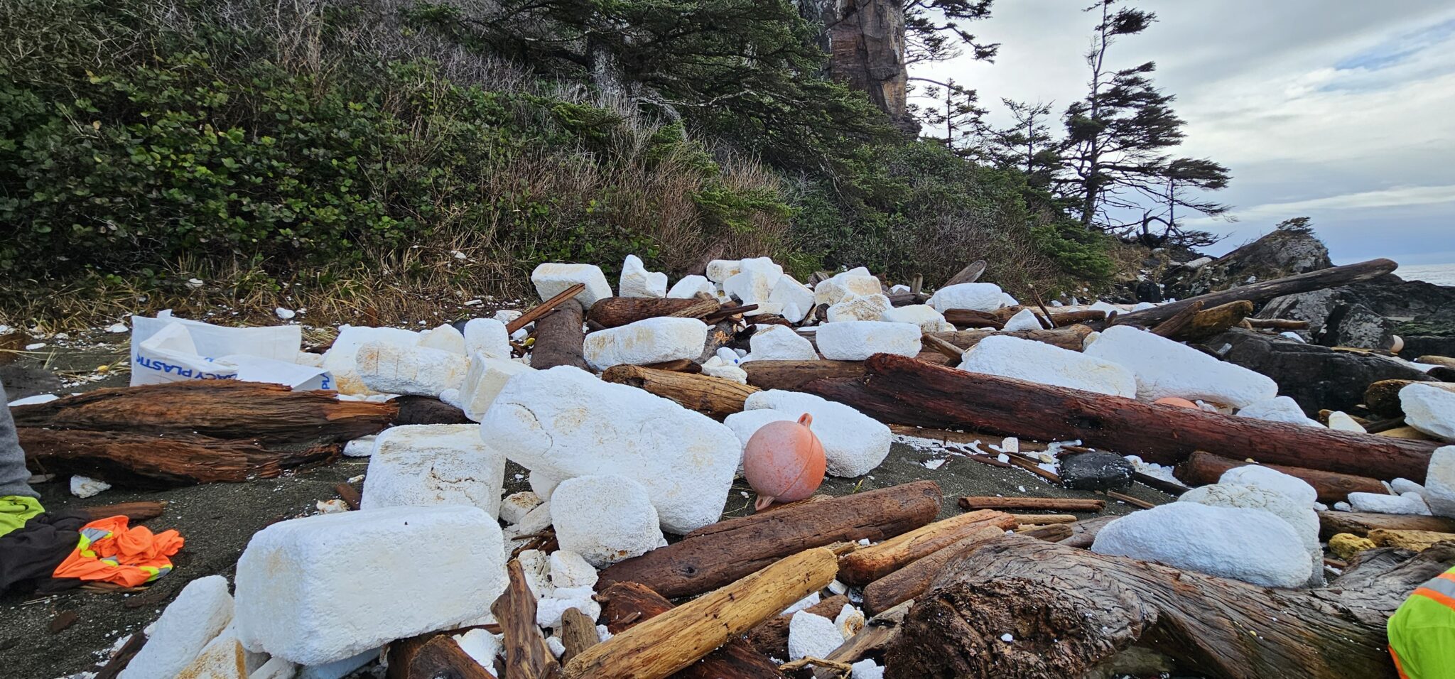 Ending Ocean Plastic Pollution with Circular Economy Solutions - remote beach with volunteers cleaning ocean plastic. Large pieces of white and yellowed styrofoam and white supersacks with Ocean Legacy's logo with buoys interspersed in driftwood and rock with additional ocean plastic on the shoreline